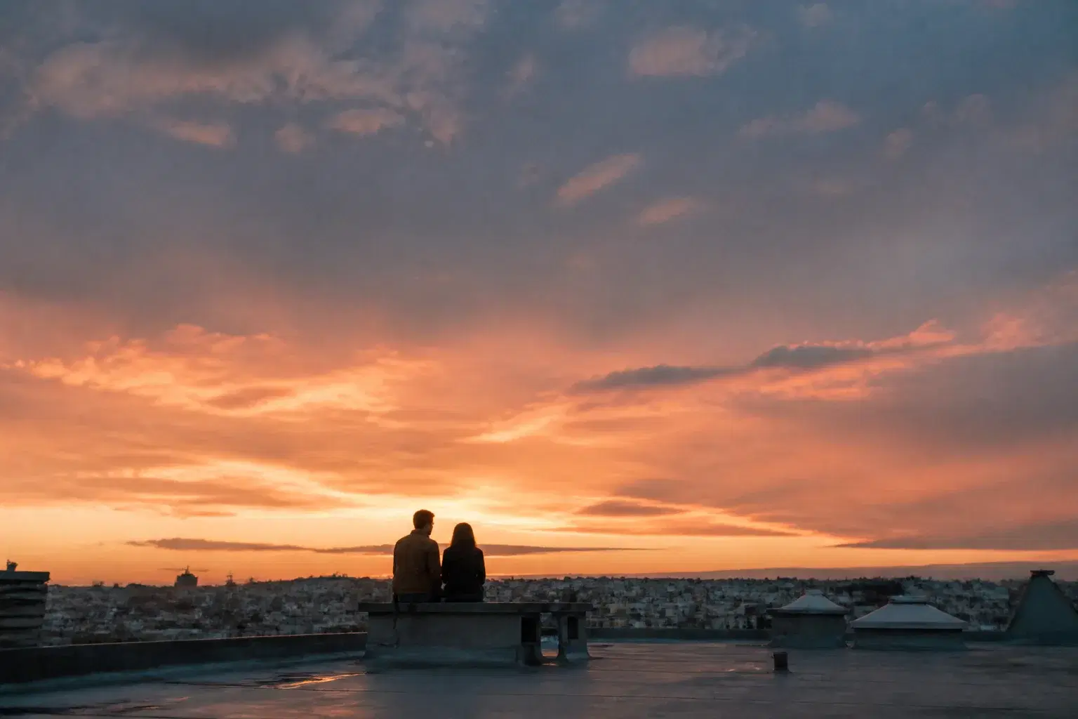 Wide cinematic view of an adult couple sitting together on a rooftop at sunset